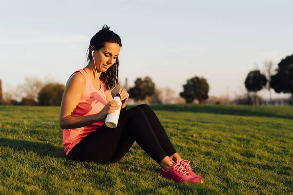 Young runner woman seated in the grass before run in a park, drinking water with smartphone and earphones. Close up athletic and healthy girl wearing white and pink sneakers.