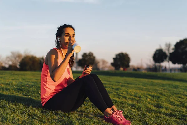 Młoda biegaczka siedząca na trawie przed bieganiem po parku, pijąca wodę ze smartfonem i słuchawkami. Zbliżenie sportowe i zdrowe dziewczyny w białe i różowe trampki. — Zdjęcie stockowe