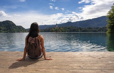 Rıhtımda oturan Bled gölünde Varsayım kilisenin panoramik manzarasının keyfini bir sırt çantası ile Gezgin kadın. En ünlü Sloven gölü ve adası Assumptio Hac Kilisesi ile Bled