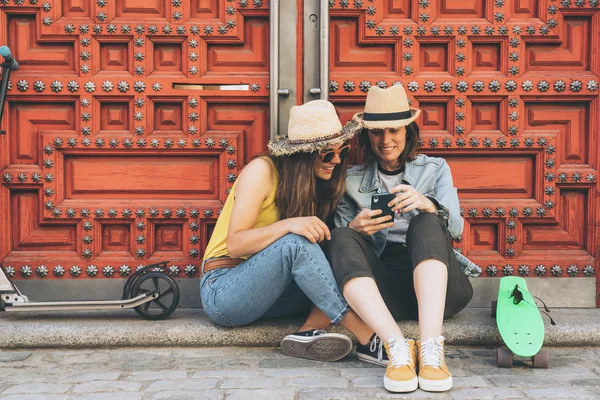 Attraktive und coole Frauen lesbisches Paar, das Handy schaut und einander vor rotem Türhintergrund anlächelt. Gleichgeschlechtliches Glück und freudiges Konzept. — Stockfoto