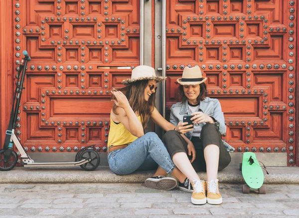 Donne attraenti e cool coppia lesbica in cerca di telefono cellulare e sorridente l'un l'altro in uno sfondo porta rossa. Felicità dello stesso sesso e concetto gioioso . — Foto Stock