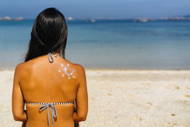 Kumsalda bronzlaşmış bir kadının omzuna güneş kremi sürmüş bir güneş çizimi.
