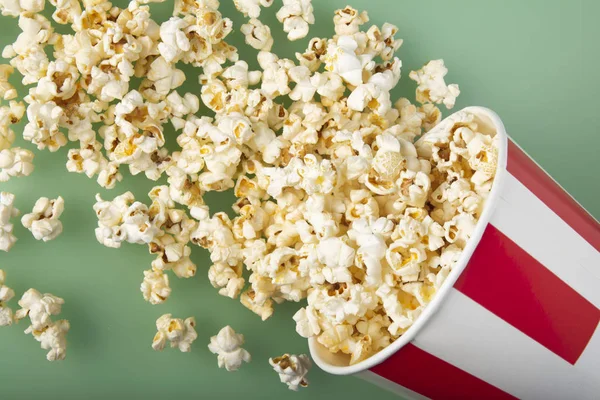 Vista superior de la taza de papel rayado volcado con deliciosas palomitas frescas sobre fondo verde. El concepto de entretenimiento . —  Fotos de Stock