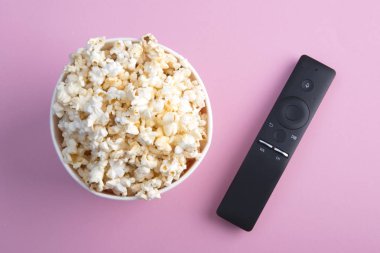Bir kase patlamış mısır, pembe arka planlı bir televizyon kumandası. Tv, film, dizi, spor, dizi izleme konsepti. Düz döşeme, üst görünüm.