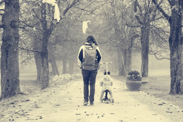 Youg mère marche avec sa fille chevauchant un scooter tout-petit dans le parc. — Photo