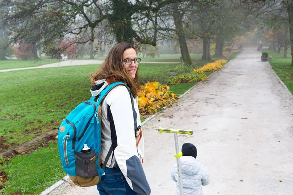 Youg mãe andando com sua filha montando uma scooter criança no parque. — Fotografia de Stock