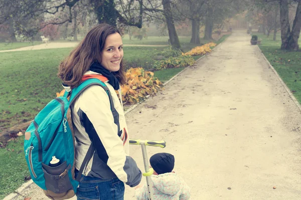 Youg madre che cammina con sua figlia cavalcando uno scooter bambino nel parco. — Foto Stock