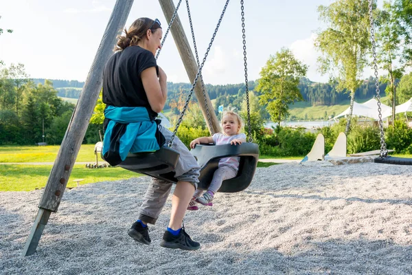 Matka a dcera houpající se na hřišti v místním parku. — Stock fotografie