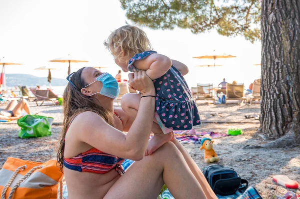 Young mother wearing medical mask playing with her daughter at the beach. New normal concept.