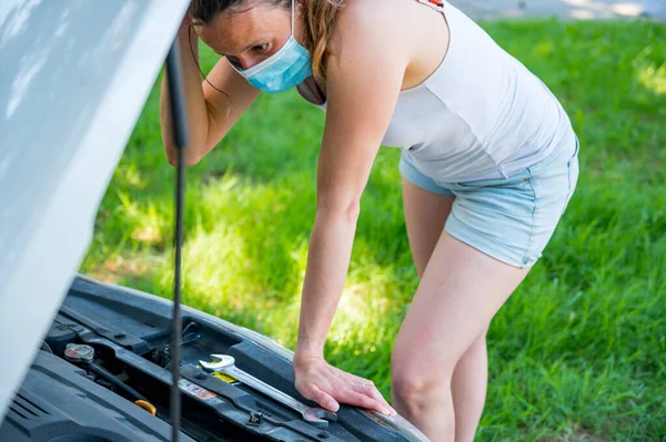 Donna che indossa maschera medica cercando di risolvere rotto motore auto. — Foto Stock