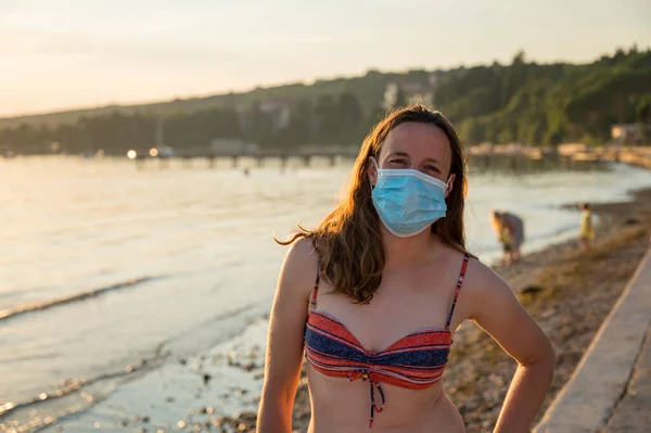 Młoda kobieta w masce medycznej na plaży podczas zachodu słońca. — Zdjęcie stockowe
