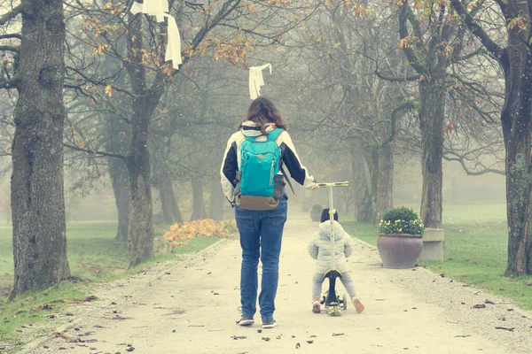 Youg mãe andando com sua filha montando uma scooter criança no parque. — Fotografia de Stock