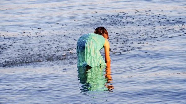 A girl stands up to her knees in the Atlantic, she bends down and plays with her hands in the water in back view. — Stock Photo, Image