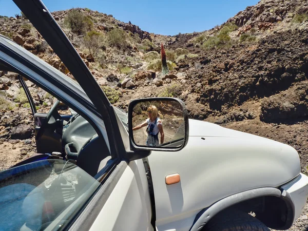 Partial view of a big car, in the right side mirror is a woman to see in the background the barren desert