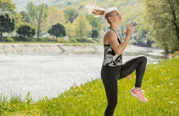 Blonde fit woman doing her training in the nature by the river. Knee-up jump. — Stock Photo, Image