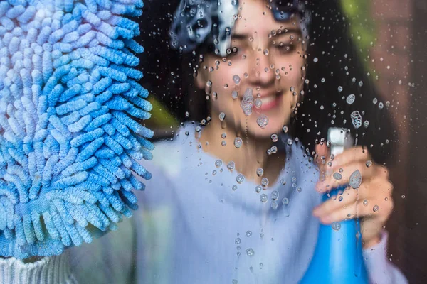 Söt flicka rengöra fönstret med blå trasa. Utsikt från utsidan. — Stockfoto