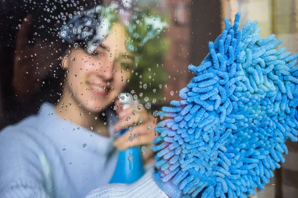 Linda chica con sonrisa en su cara limpiando la ventana con trapo azul . — Foto de Stock