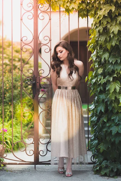 Beautiful teen girl in glamorous golden dress standing by the gate. — Stock Photo, Image