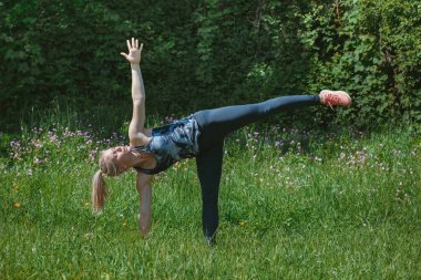 Sarışın kız doğada tek bacaklı aşağı köpek yoga poz yapıyor.