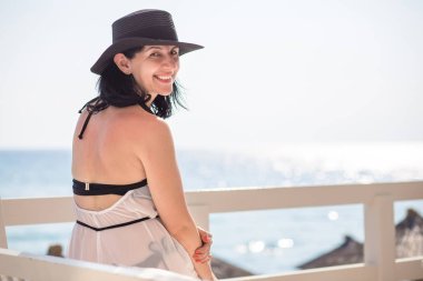 Siyah şapkalı kadın, ahşap rıhtımda mayo yla ayakta, gülümseyerek.