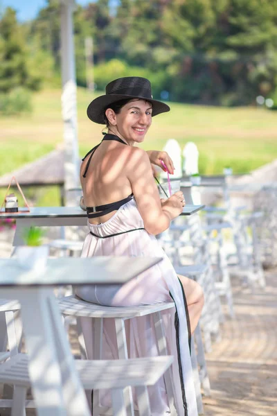 Woman with black hat and white dress, sitting in sea bar, drinking cocktail.