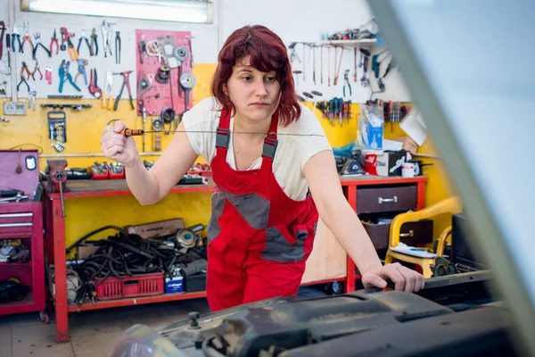 Female mechanic in red work wear overalls in car mechanic shop checking car oil.