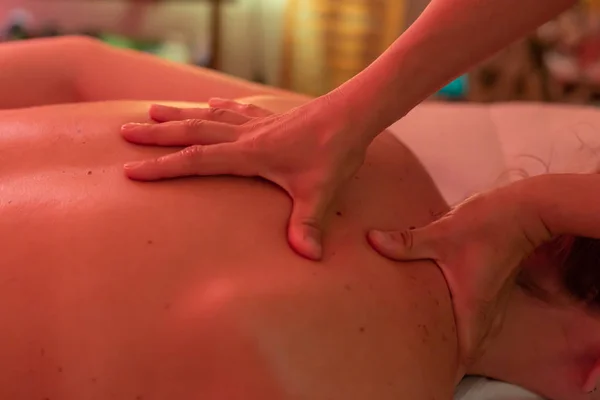Close-up photo of masseuse massaging her client back and neck. Red spa light.