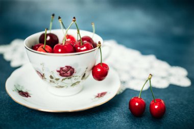 Masanın üzerinde, altında tığ işi olan porselen bir fincanda kırmızı, lezzetli kirazlar. Mavi arkaplan