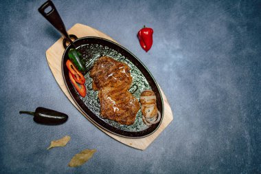 Bir parça ızgara biftek, yanında kırmızı biber ve soğan. Siyah tavada, mavi arka planda. Üst görünüm