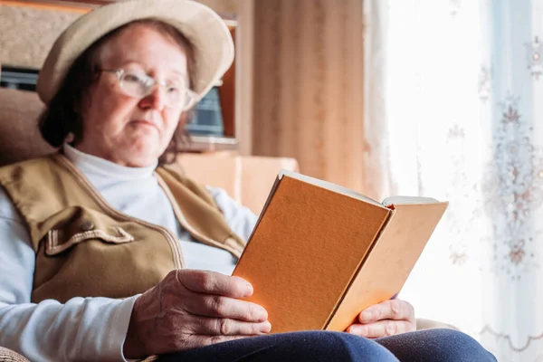 stock image Elderly woman with a hat on, reading a book by the window. Afternoon light