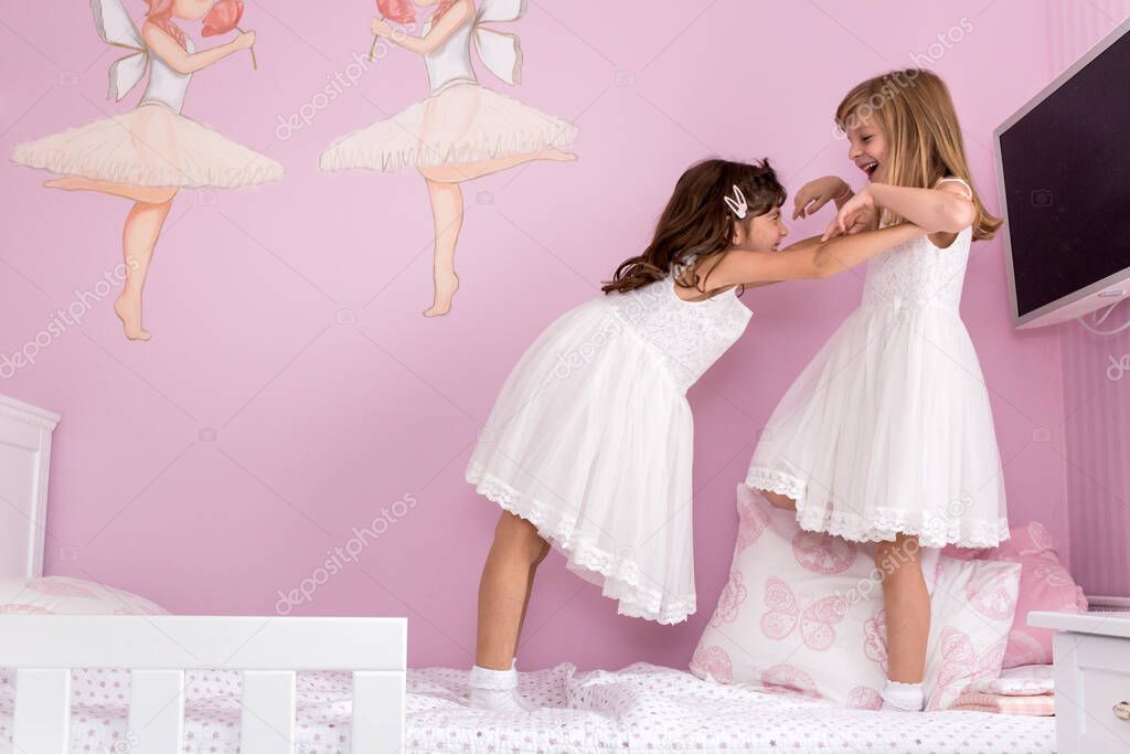Adorable, cute, little girls, sisters, twins playing on the bed in their lovely pink room in white dresses