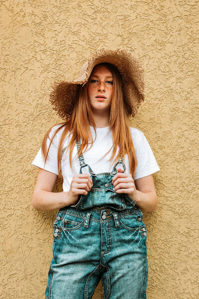 Portrait of young, teen ginger girl in blue jeans with suspenders and a hat. Yellow backgound