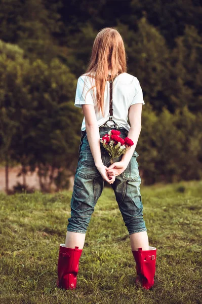 Back view photo of ginger girl in jeans with suspenders and red boots holding bouquet of roses behind her back