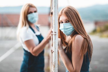 Anne ve kızı çitin farklı taraflarında durmuş tıbbi maskeler takmış kameraya bakıyor, kıza odaklanıyor. Sosyal uzaklık kavramı