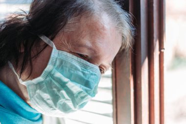 Yüzünde cerrahi maske olan yaşlı bir kadının dışarıdaki pencereden dışarı bakarken çekilmiş fotoğrafı.