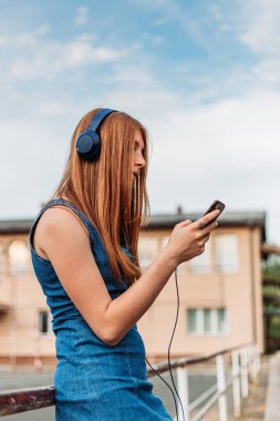 Hoş, kızıl kız telefonunda müzik çalıyor. Kulaklıkları kafasında, okul bahçesinde.