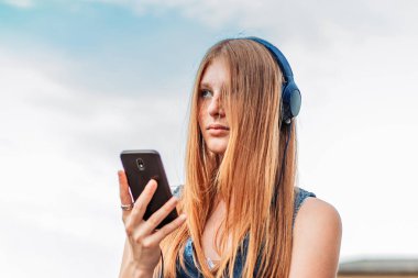 Hoş, kızıl kız telefonunda müzik çalıyor. Kulaklıkları kafasında, okul bahçesinde.