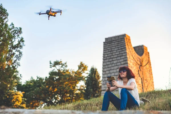 Young woman flying drone in the park, sitting on the ground, looking at it