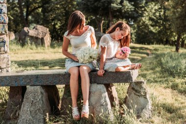 Genç anne ellerinde buketlerle parkta kızıyla oturuyor.