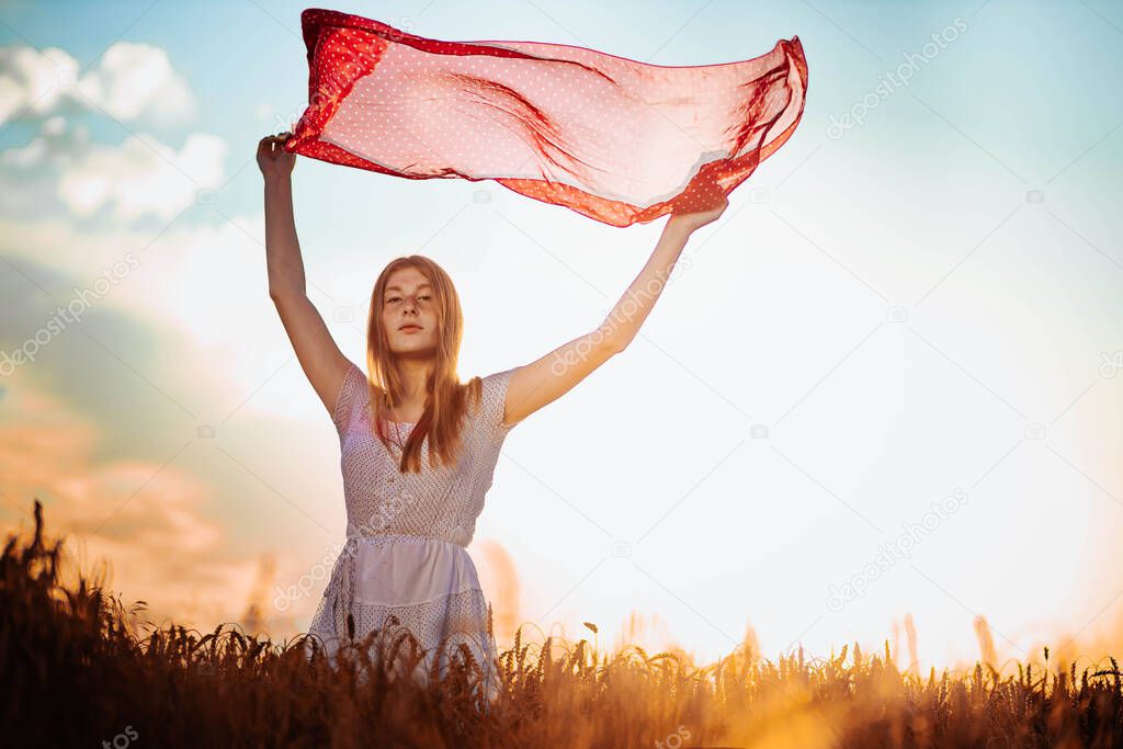 Outdoors photo of young, ginger girl in white dress holding a red scarf fluttering up in the air. Copy space