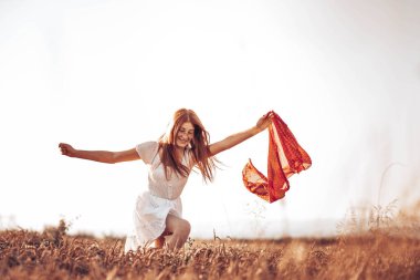 Buğday tarlasında elinde kırmızı bir atkıyla koşan beyaz elbiseli, kızıl saçlı genç bir kızın ön görüntüsü. Boşluğu kopyala