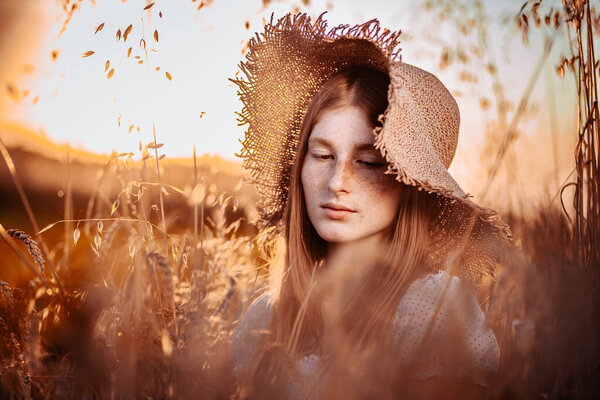 Close-up portrait of young, teen, ginger girl with freckles wearing a summer hat. In the wheat field