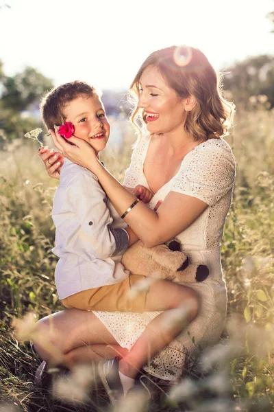 美しい若いお母さんが息子を抱いて耳の後ろにバラをつけた 屋外の写真 母の日 — ストック写真