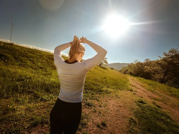 Arka planda spor giyimli genç sarışın bir kızın at kuyruğu bağladığı bir fotoğraf var. Sabah ışığı. Güneşli bir hava. Sağlıklı yaşam tarzı ve spor anlayışı