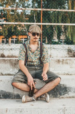 Retro giysili genç hippi merdivenlerde elinde birayla oturuyor.