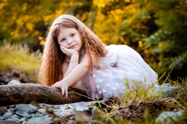 Beyaz elbiseli şirin, kızıl kız bir tahta parçasına yaslanmış, fotoğraf için poz veriyor.