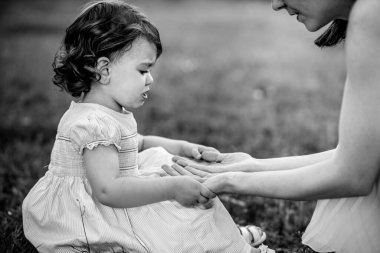 Siyah beyaz bir portre, üzgün görünen küçük, kıvırcık bir kız, annesini teselli etmek için elini tutuyor.