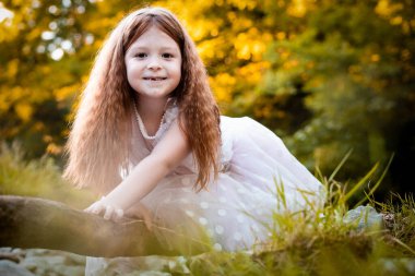 Sevimli, küçük, kızıl saçlı, beyaz elbiseli kız bir tahta parçasına yaslanmış, fotoğraf için poz veriyor.