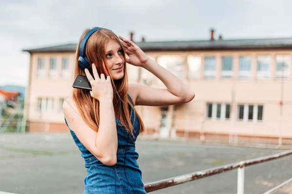 Cute, ginger girl streaming music on her phone. Headphones on her head, school yard