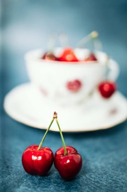 Masadaki eski bir porselen bardağın önünde iki kırmızı, lezzetli kiraz. Mavi arkaplan
